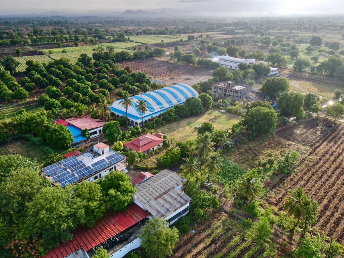 The campus from the air: red-tiled roofs, solar panels, the blue-striped dome of Shanti Kutir, mango orchards and ploughed fields reaching to the hills.