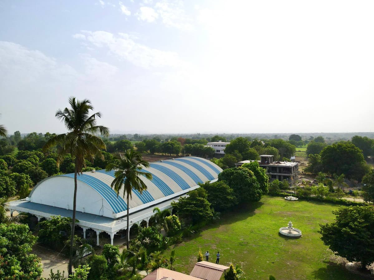 Shanti Kutir from above — a long white hall with a blue-and-white striped barrel roof among palms, beside a green lawn with a fountain.
