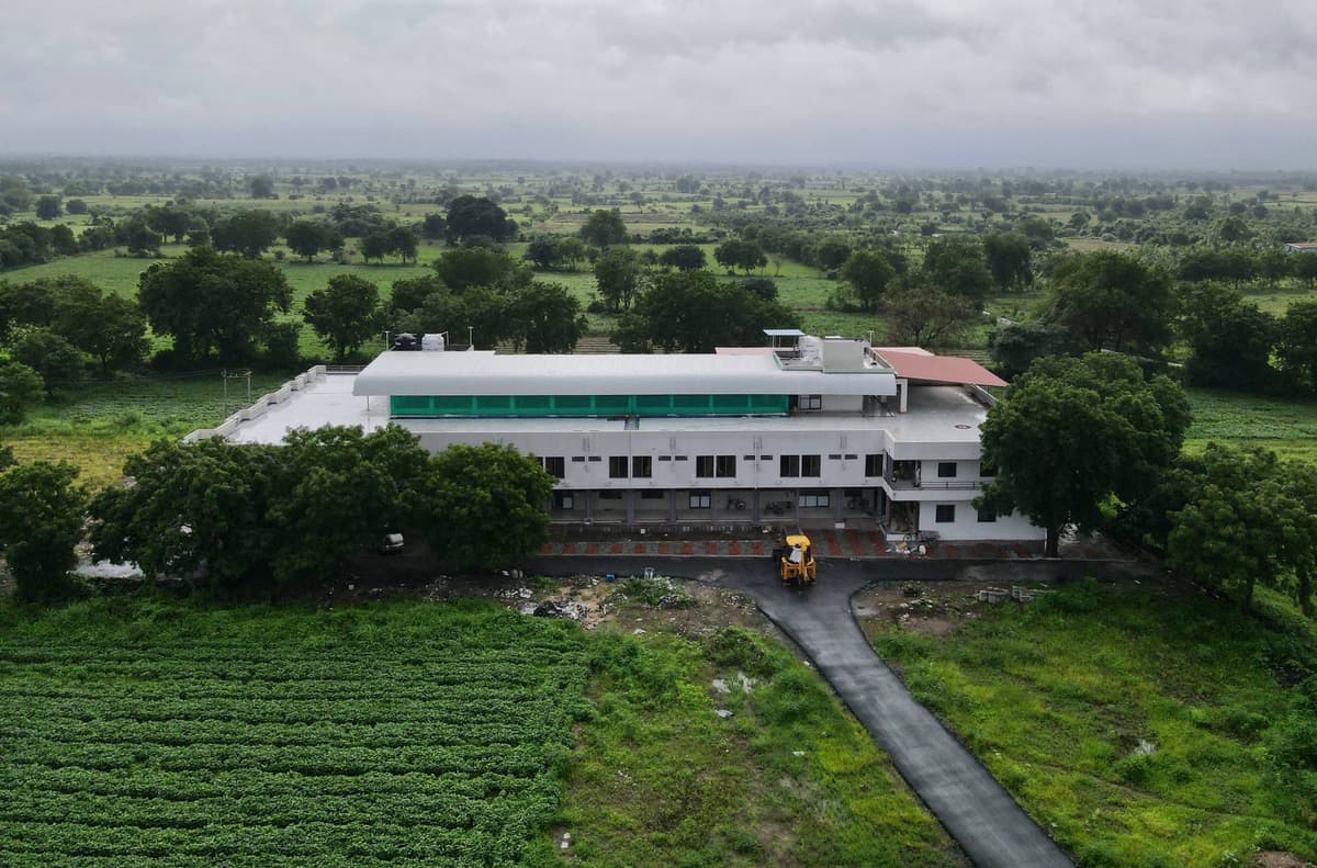The white building under grey monsoon clouds, a curved black road leading to it, green crops in front and a yellow digger at the door.