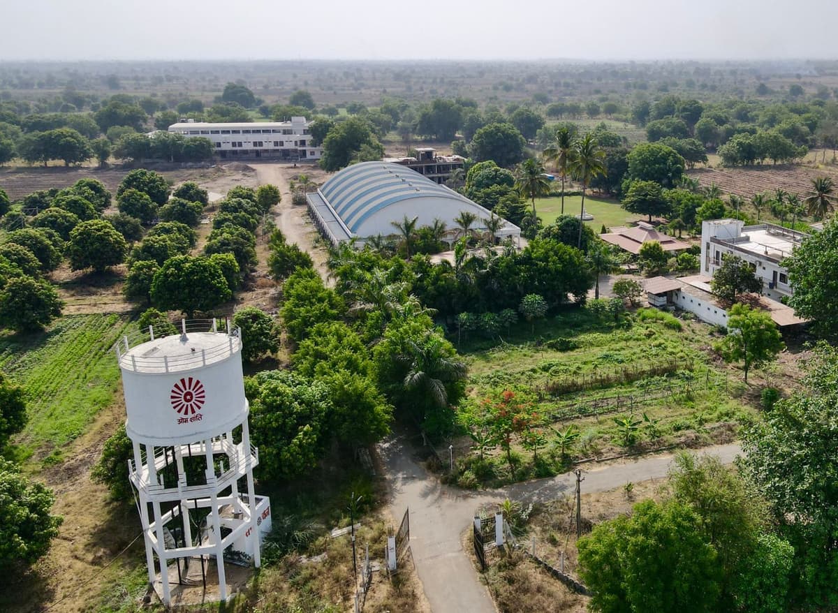 A white water tower marked “Om Shanti” at the gate, with the striped dome, mango rows and a white building behind.